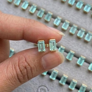 May include: A pair of gold stud earrings with rectangular, light blue gemstones. The earrings are held in a hand, with several other pairs of earrings visible in a gray jewelry tray. The jewelry is from the brand "JEWELNRY".