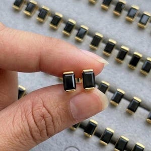 Puede incluir: Pendientes de botón dorados con piedras negras rectangulares. Los pendientes se sostienen en una mano, con otros pendientes similares dispuestos en filas sobre una superficie gris. Marca: "JEWENLERY JEWELRY".