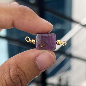 May include: A small, square, deep purple gemstone bead with gold-colored loops on either side. The gemstone has a slightly rough texture and is held between two fingers against a blurred background.