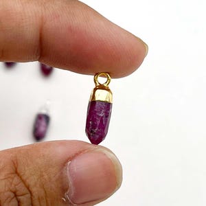 May include: A small, faceted ruby pendant with a gold-colored cap and loop. The ruby is a deep, rich red color. The pendant is held between two fingers against a white background. Several other pendants are visible in the background.