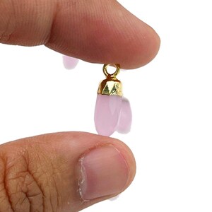 May include: A small, light pink charm with a gold-colored cap and loop. The charm is held between two fingers against a white background. The charm has a faceted, teardrop shape.