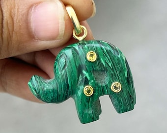 Könnte beinhalten: Ein grüner Malachit-Elefantenanhänger mit goldfarbenen Akzenten. Der Anhänger zeigt ein geschnitztes Elefanten-Design mit drei gelbgoldfarbenen Verzierungen. Der Anhänger ist an einer goldfarbenen Öse aufgehängt.