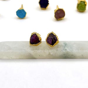 Peut inclure: Une collection de boucles d'oreilles à clous en pierres précieuses avec des montures dorées. Les boucles d'oreilles présentent diverses couleurs, notamment vert, bleu, violet et rose. Deux boucles d'oreilles sont présentées sur une base rectangulaire blanche.