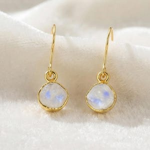 May include: A pair of gold drop earrings featuring small, round, translucent moonstones with a subtle blue iridescent sheen. Each stone is encased in a smooth gold bezel setting and hangs from a simple, curved gold ear wire hook.