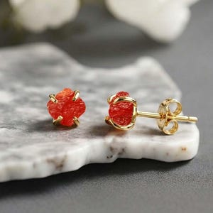 May include: A pair of stud earrings featuring vibrant red gemstones set in gold-colored metal. The gemstones have a rough, natural appearance. The earrings are displayed on a light gray surface, with white flowers in the background.