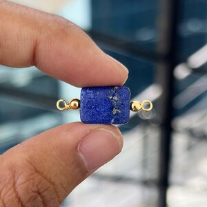 Può includere: Una piccola perla rettangolare di lapislazzuli blu intenso con dettagli color oro. La perla è tenuta tra due dita, mostrando il suo ricco colore e la sua consistenza. I dettagli dorati sembrano essere piccoli anelli.