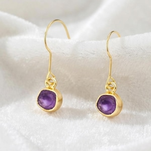 May include: A pair of gold-toned drop earrings featuring small, round, faceted purple amethyst stones set in smooth metal bezels. Each earring hangs from a simple, curved gold-toned hook wire, showcasing a minimalist design with a polished finish.