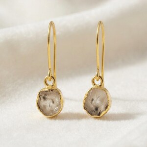 May include: A pair of gold drop earrings featuring small, raw, translucent grey stones with irregular, organic shapes. Each stone is encased in a thin gold bezel setting and hangs from a simple, curved gold ear wire hook for a clean, minimalist look.