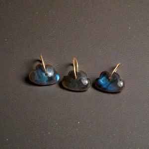 May include: Three cloud-shaped earrings with gold-coloured hooks. The earrings are made of a dark, iridescent stone with blue and green flashes. The earrings are displayed on a dark grey surface.