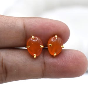 May include: A pair of orange gemstone stud earrings. Each earring features an oval-shaped, translucent orange stone set in a gold-colored metal frame. The earrings are held in a hand against a white background.