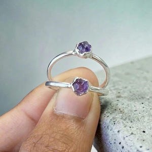 May include: Two silver rings, each with a raw, faceted purple amethyst gemstone. The rings are set against a soft grey background, highlighting the minimalist design and natural beauty of the stones.