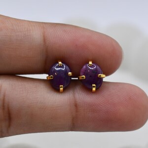 May include: A pair of oval-shaped purple gemstone stud earrings set in gold-toned metal prongs. Each earring features a smooth, polished cabochon-cut stone with deep violet hues, securely held by four small gold-colored metal prongs.