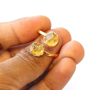May include: A gold-colored ring featuring two raw, yellow-orange citrine gemstones. The ring's band wraps around the finger, with each stone set in a gold bezel. The gemstones have a natural, irregular shape, adding to the ring's unique design.