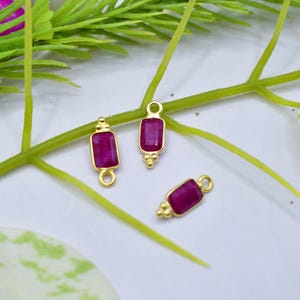 Può includere: Tre elementi di gioielleria color oro con pietre preziose rettangolari rosa intenso. Gli elementi hanno un anello nella parte superiore e dettagli decorativi. Sono disposti su una superficie bianca con steli e foglie verdi.