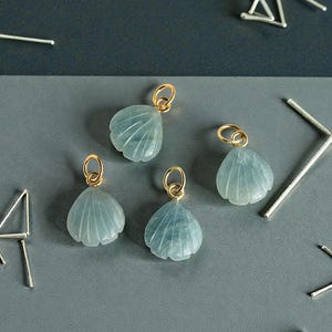 Könnte beinhalten: Vier hellblaue Muschel-Anhänger mit goldfarbenen Schlaufen. Die Anhänger sind mit detaillierten Linien geschnitzt und auf einer grau-dunkelblauen Oberfläche angeordnet. Mehrere silberfarbene geometrische Formen sind verstreut.