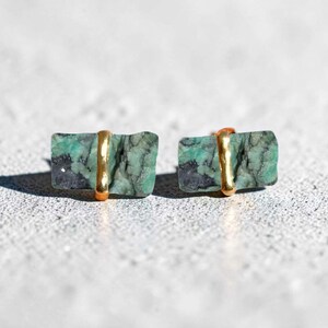 Puede incluir: Un par de pendientes de botón rectangulares. Cada pendiente presenta una piedra verde y negra de corte rugoso acentuada con una banda dorada. Los pendientes están sobre un fondo gris claro.