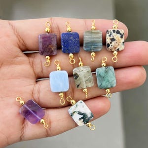 Hand holding a collection of natural crystal gemstone charms with gold bails. Includes square and rectangular pendants made of amethyst, lapis lazuli, blue lace agate, moss agate, dalmatian jasper, and other semi-precious stones, perfect for jewelry making