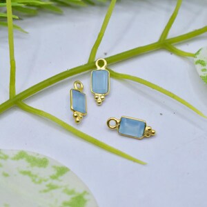 May include: Three gold-colored rectangular charms with light blue centers. Each charm has a small loop at the top and decorative gold accents at the bottom. The charms are displayed on a white surface with green plant stems.