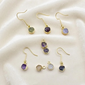 May include: A collection of gold-toned earrings and pendants featuring raw, uncut gemstones in various colors including green, purple, blue, and clear. Each piece has a metal hook or loop, showcasing natural, irregular stone shapes and textures.