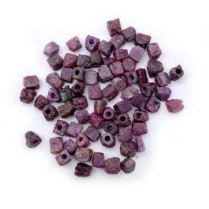 May include: A collection of raw, cube-shaped ruby beads in varying shades of red and purple. Each bead has a hole drilled through it, ready for stringing. The beads are scattered on a white surface.