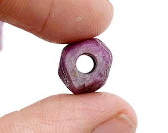 May include: A close-up of a faceted, deep red ruby bead with a hole drilled through the center, held between two fingers. Other rough ruby stones are scattered in the background against a white backdrop. The bead is approximately 0.5 inches in diameter.