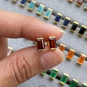 Può includere: Orecchini a bottone dorati con pietre rettangolari rosso scuro. Gli orecchini sono mostrati davanti a un vassoio di gioielli grigio pieno di orecchini simili in vari colori. Il logo Jewenlery Jewelry è visibile.