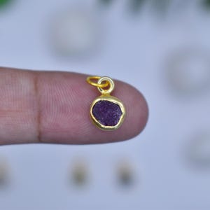 May include: A small, gold-colored pendant with a raw, deep purple gemstone. The pendant has a simple, circular design with a loop at the top for a chain. The gemstone is set in a gold bezel, creating a minimalist and elegant look.