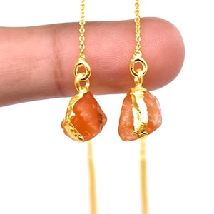 Puede incluir: Un par de pendientes dorados con piedras preciosas en bruto de color naranja. Las piedras están engastadas en metal dorado y cuelgan de finas cadenas doradas. Los pendientes se muestran sobre un fondo blanco.
