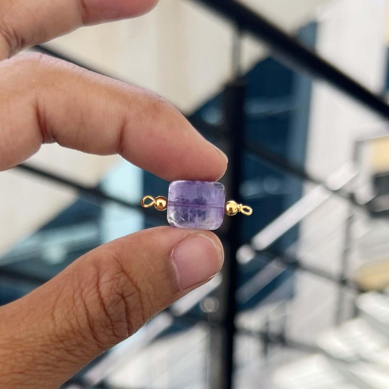 Close-up of fingers holding a single natural amethyst gemstone charm with gold bail. The square purple crystal pendant shows beautiful translucent layers and natural inclusions, ideal for handmade jewelry making.