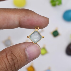 May include: A square-shaped charm featuring a faceted light blue gemstone set in a gold-toned metal frame. The charm includes a small circular loop at the top for attachment and two decorative metal accents on the left and right sides of the setting.