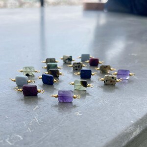 Assortment of natural crystal gemstone charms with gold bails arranged on a smooth gray surface. Square and rectangular pendants featuring amethyst, lapis lazuli, blue lace agate, moss agate, dalmatian jasper, ruby in zoisite, and other semi-precious stones, perfect for handmade jewelry making.