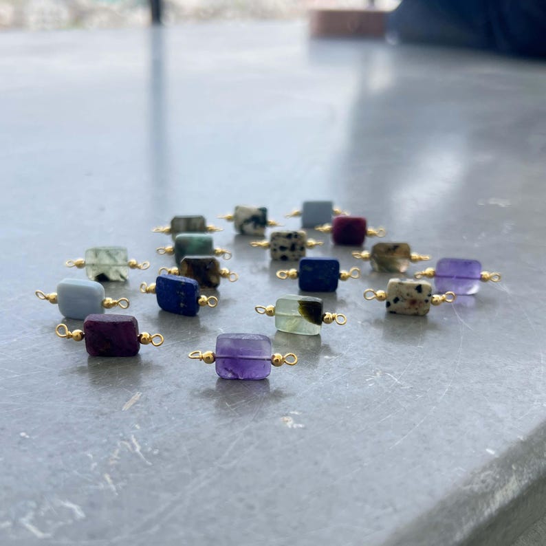 Assortment of natural crystal gemstone charms with gold bails arranged on a smooth gray surface. Square and rectangular pendants featuring amethyst, lapis lazuli, blue lace agate, moss agate, dalmatian jasper, ruby in zoisite, and other semi-precious stones, perfect for handmade jewelry making.