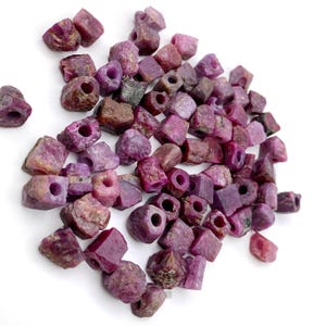 May include: A collection of raw ruby gemstone beads in various shapes and sizes. The beads are primarily shades of deep red and purple, with visible natural textures and drilled holes for stringing. The background is plain white, highlighting the gemstones.