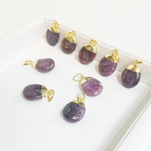 May include: A collection of oval-shaped pendants with a gold-colored cap and bail. The pendants are a mix of purple and maroon hues, and are arranged on a white surface. The gold caps add a touch of elegance to the natural stone pendants.