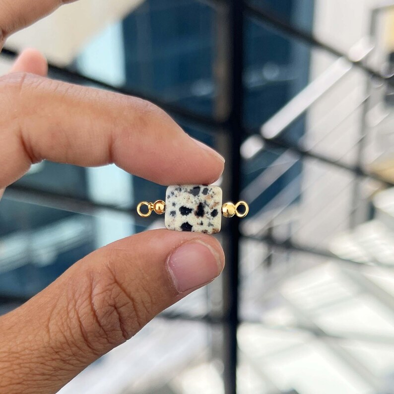 Close-up of fingers holding a single natural dalmatian jasper gemstone charm with gold bail. The square white pendant features distinctive black and brown spots, perfect for handmade jewelry making.