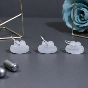 May include: Three cloud-shaped earrings with silver hoops. The earrings are made of a translucent, light-coloured stone. The earrings are displayed on a grey surface with a gold wire structure and a blue flower in the background.