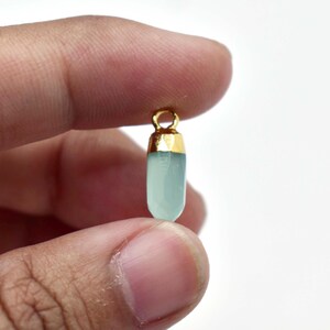 May include: A small, light blue gemstone pendant with a gold-colored cap and loop. The pendant is held between fingers against a white background. The gemstone has a faceted, elongated shape.