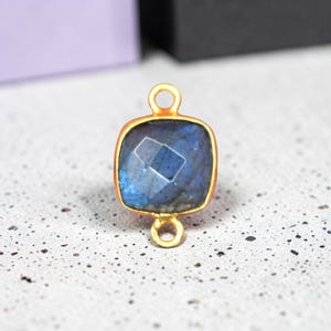 Könnte beinhalten: Ein quadratischer Labradorit-Edelsteinverbinder mit facettierter Oberfläche in blauen und grauen irisierenden Tönen. Der Stein ist in eine goldfarbene Metallfassung eingefasst und verfügt über eine kleine runde Öse oben und unten zur Befestigung.