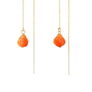 May include: A pair of gold-colored threader earrings. Each earring features a small, irregular orange stone suspended from a delicate chain. A straight, gold-colored bar hangs below the stone, creating a simple yet elegant design.