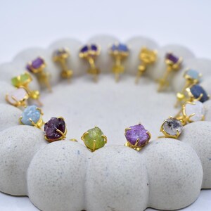 May include: A collection of gemstone stud earrings in various colors, including purple, green, blue, and pink. Each earring features a raw, uncut gemstone set in a gold-colored metal setting. The earrings are arranged in a circular pattern.