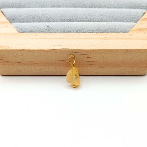 May include: A small, gold-colored pendant featuring a rough-cut, light-yellow gemstone. The pendant is suspended from a delicate gold loop. The gemstone has a natural, irregular shape. The pendant is displayed against a wooden background.