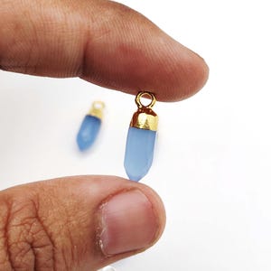 May include: Close-up of two light blue crystal pendants with gold-colored caps. The pendants are held between fingers against a white background. The crystals have a faceted design.