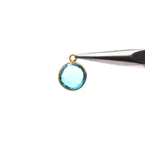 May include: A round, light blue gemstone pendant with a gold-colored bezel and loop. The pendant is held by a pair of silver tweezers against a white background. The gemstone has a clear, polished appearance.