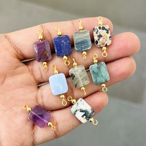 Hand holding a beautiful collection of natural crystal gemstone charms with gold bails. Square and rectangular pendants made from amethyst, lapis lazuli, labradorite, dalmatian jasper, green moss agate, blue lace agate, phantom quartz, and other semi-precious stones – perfect for jewelry making.