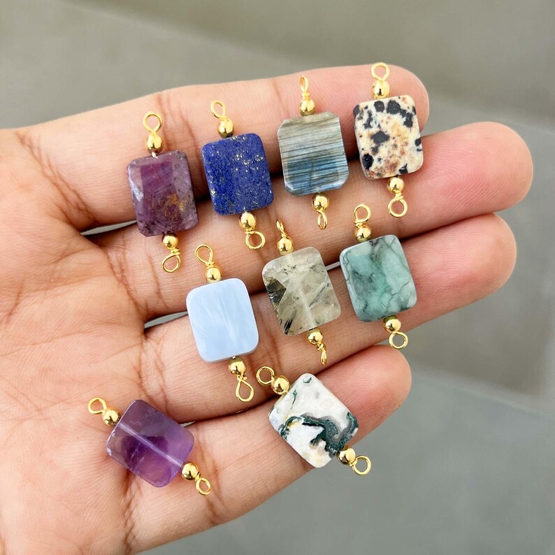 Hand holding a beautiful collection of natural crystal gemstone charms with gold bails. Square and rectangular pendants made from amethyst, lapis lazuli, labradorite, dalmatian jasper, green moss agate, blue lace agate, phantom quartz, and other semi-precious stones – perfect for jewelry making.