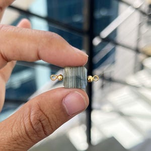 Close-up of fingers holding a single natural labradorite gemstone charm with gold bail. The square slice pendant shows beautiful striped and iridescent patterns, perfect for handmade jewelry making.