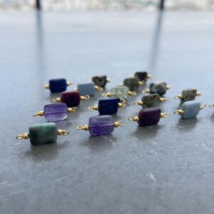 Collection of natural crystal gemstone charms with gold bails arranged on a reflective gray surface. Square and rectangular pendants made from amethyst, lapis lazuli, green moss agate, purple ruby in zoisite, blue lace agate, and other semi-precious stones, perfect for handmade jewelry making