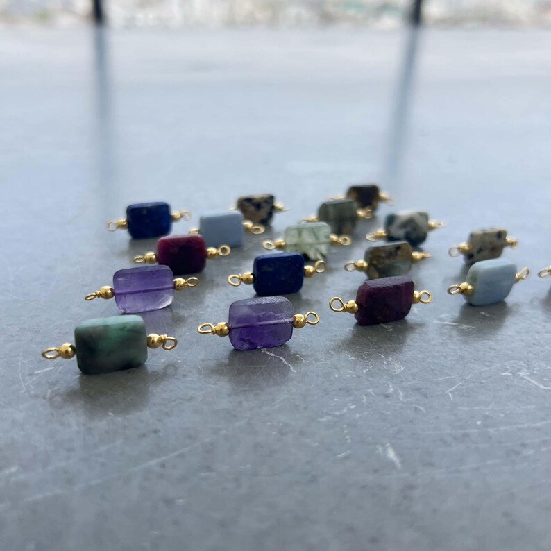 Collection of natural crystal gemstone charms with gold bails arranged on a reflective gray surface. Square and rectangular pendants made from amethyst, lapis lazuli, green moss agate, purple ruby in zoisite, blue lace agate, and other semi-precious stones, perfect for handmade jewelry making