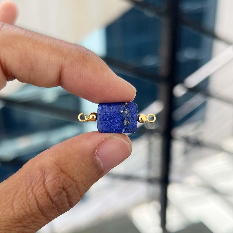 Close-up of fingers holding a single natural lapis lazuli gemstone charm with gold bail. The vibrant blue square pendant shows beautiful natural golden pyrite flecks, perfect for handmade jewelry making.|
Natural Lapis Lazuli Slice Charm with Gold Bail – Close-up of a rich blue lapis lazuli rectangular pendant with golden pyrite inclusions, held between fingers for jewelry making.
Single lapis lazuli crystal charm with gold bail held between fingers.