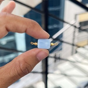 Close-up of fingers holding a single natural blue lace agate gemstone charm with gold bail. The soft light blue square pendant displays delicate white banding patterns, perfect for handmade jewelry making.
Hand holding a natural blue lace agate square gemstone pendant with gold bail.
Natural Blue Lace Agate Slice Charm with Gold Bail – Close-up of a soft light blue agate rectangular pendant with white banding, held between fingers for jewelry making.